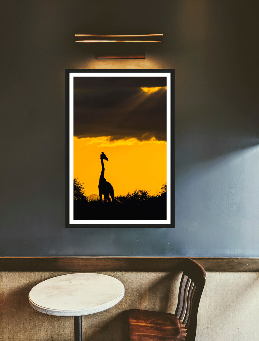 A photograph of a giraffe silhouette standing under a stormy orange sky during sunset hanging on a greyish wall above a table & chair.