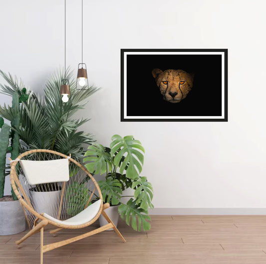 A photograph showcasing the amber eyes and a Cheetah's face up close appearing from the darkness hanging on a white wall in a room with a chair and plants next to it.