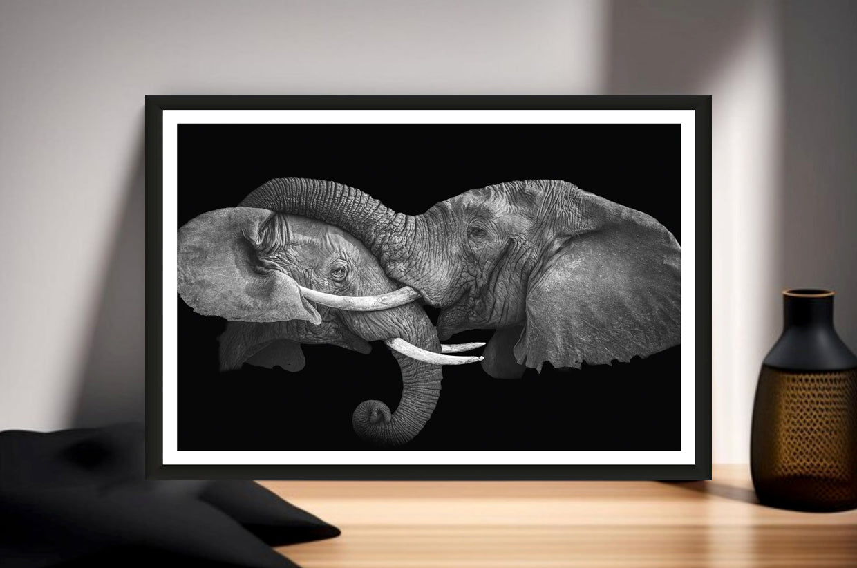 Photograph of one elephant having his trunk on the head of another elephant standing on a sideboard next to a vase.