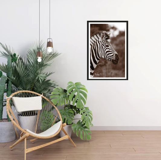 A photograph of a zebra's head in sepia colors with a blurred background hanging on a white wall in a room with a chair and plants next to it.