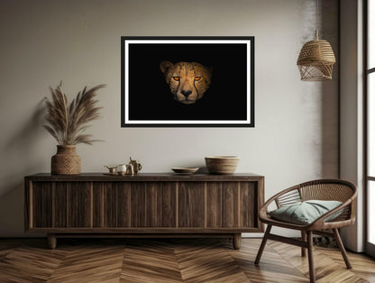 A photograph showcasing the amber eyes and a Cheetah's face up close appearing from the darkness hanging on a brown wall above a chair and a commode.